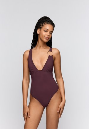 Vrouw met lang gevlochten haar die een diep bordeauxrode badpak draagt met een metalen ringdetail op de linkerschouder.