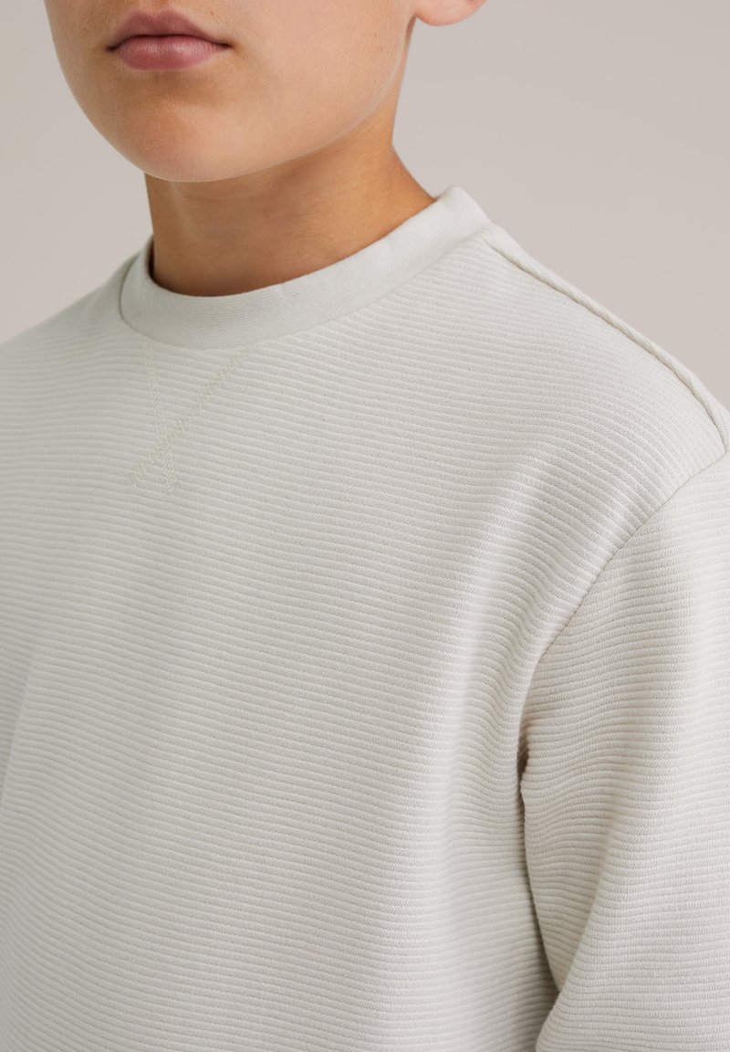 Gai pelēks, ribots džemperis ar apaļu izgriezumu, kuram ir smalkas šuves un mīksta tekstūra. Dizains ir minimāls un mūsdienīgs.