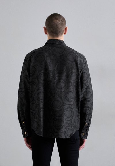 Chemise noire à motif avec une coupe décontractée, dotée d'un col et de accents en boutons dorés au niveau des poignets. Tissu texturé.
