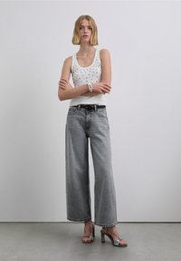 Bílý tílko bez rukávů se stříbrnými cvoky, volné šedé džínové kalhoty, černý pásek a stříbrné sandály na vysokém podpatku se stříbrnými pásky. Minimalistický design.