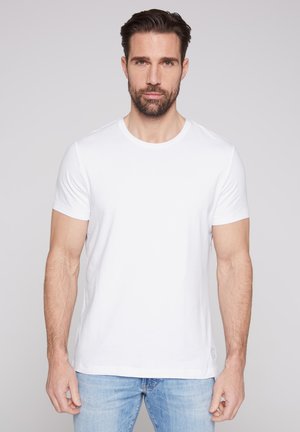 Mann mit kurzen dunklen Haaren und Bart, der ein schlichtes weißes kurzärmeliges T-Shirt und hellblaue Jeans trägt, steht vor einem grauen Hintergrund.