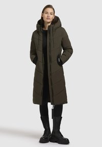 Dlouhá olivově zelená péřová bunda s kapucí, diagonálně prošívaný vzor, zipové zapínání a boční kapsy. V kombinaci s černými šněrovacími bojovými botami.