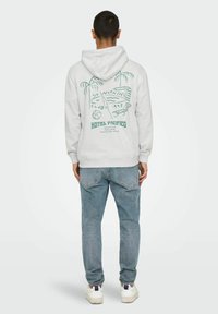 Sweat à capuche gris en mélange de coton, avec un imprimé graphique vert représentant des palmiers et du texte au dos. Associé avec un jean bleu clair et des baskets.