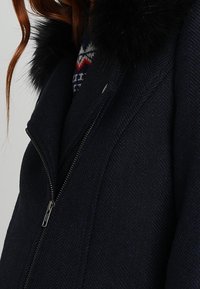 Närbild av en mörk marinblå kappa med dragkedja framtill och svart konstpälskrage, bärs över en mönstrad tröja.