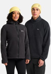 Zwei Personen, die schwarze Fleecejacken und gelbe Strickmützen tragen, stehen nebeneinander vor einem weißen Hintergrund.