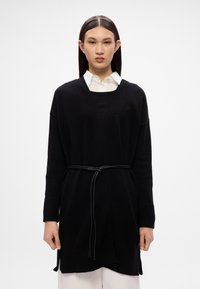 Μαύρο πλεκτό cardigan με βαθύ V λαιμό, ριγκσμάν, πλαϊνές σχισμές και μια μαύρη ζώνη που τονίζει τη μέση, στρωμένο πάνω από λευκό πουκάμισο.