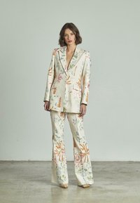 Donna in piedi che indossa un abito color crema con motivi floreali colorati, pantaloni a gamba larga e un lungo blazer contro uno sfondo semplice.