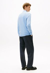 Młody mężczyzna w jasnoniebieskim swetrze i ciemnych spodniach stoi z rękami w kieszeniach, lekko odwracając się w stronę białego tła.