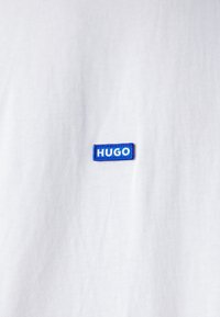 T-shirt bianco in cotone con una patch rettangolare blu con il testo "HUGO" in bianco. Texture liscia con una vestibilità classica.