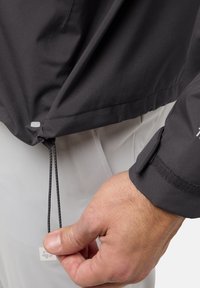 Sort vandtæt jakke med en glat tekstur. Har en justerbar kant og velcro-fremhævede håndled. Nærbillede af en hånd, der justerer snoren.