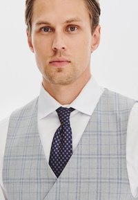Szary kamizelka w kratę na białej koszuli, zestawiona z granatowym krawatem w geometryczny wzór. Gładka faktura materiału, dopasowany krój.