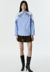 Camicia azzurro chiaro con pannelli a strisce sulle spalle e intricate ricami neri. Abbinata a una gonna plissettata marrone e stivali neri al ginocchio.