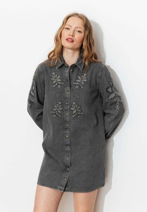 Femme blonde portant une robe chemise en jean noire avec une broderie florale, debout contre un fond blanc, les mains derrière le dos.
