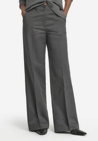 Weite Hose aus dunkelgrauem Stoff mit glatter Textur, vorderen Taschen und einem Knopfverschluss. Schwarze spitze Schuhe runden den Look ab.