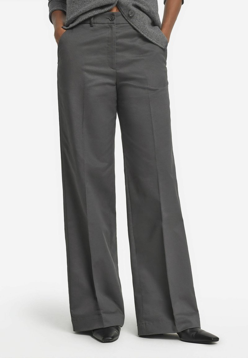 Weite Hose aus dunkelgrauem Stoff mit glatter Textur, vorderen Taschen und einem Knopfverschluss. Schwarze spitze Schuhe runden den Look ab.
