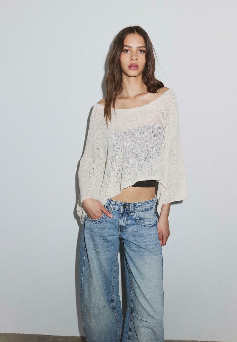 Žena nosi voľný smotanovo biely sveter cez čierny crop top a svetlomodré džíny s širokými nohavicami, stojaca pred jednoduchou svetlosivou stenou.