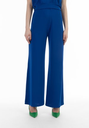 Frau trägt hoch taillierte, weit geschnittene blaue Hose, kombiniert mit grünen spitzen Schuhen, steht vor weißem Hintergrund.