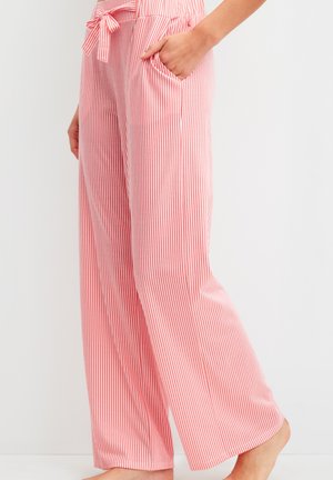 Vrouw die een broek met hoge taille, wijde pijpen en verticale rood-witte strepen draagt, met een gestrikte tailleband, hand in de zak, op blote voeten.