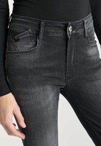 Jean en denim noir taille haute avec une texture légèrement délavée, dotée d'une poche avant, de passants de ceinture et d'une fermeture à bouton visible.