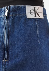 Närbild av blå denim Calvin Klein-jeans med synlig dragkedja och sytt vitt logomärke på midjan.