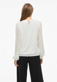 Blusa color crema con maniche lunghe e trasparenti, scollo rotondo e dettaglio a chiave sul retro, abbinata a pantaloni neri a gamba larga.