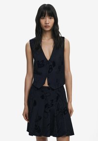 Gilet marine avec un décolleté en V profond et des boutons à l'avant, associé à une jupe plissée assortie avec un motif floral subtil. Texture du tissu lisse.