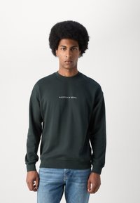 Sweatshirt vert foncé avec des poignets et une taille côtelés, arborant un logo blanc "SCOTCH & SODA" sur la poitrine, fabriqué à partir d'un matériau doux.