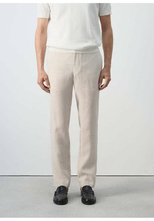Mann, der beige Leinenhose, weißes kurzärmeliges Hemd und schwarze Lederloafers trägt, steht vor einem einfarbigen hellen Hintergrund.