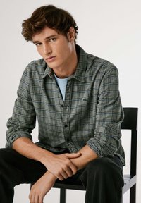 Camicia di flanella a quadri grigi con accenti blu e neri, frontale con bottoni, vestibilità comoda, indossata sopra una camicia azzurra chiara, abbinata a pantaloni scuri.