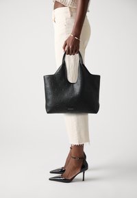 Sac cabas en cuir noir embossé avec deux poignées et une plaque de marque. Portez-le avec un pantalon beige court et des talons aiguilles noirs.
