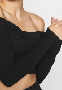 Black ribbed off-the-shoulder top with long sleeves, featuring a fitted design and soft texture; cropped length highlights the waist.