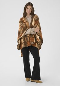 Poncho à motifs marron et orange avec un tricot texturé, présentant des manches larges et une bordure festonnée, porté sur un chemisier beige et un pantalon noir.