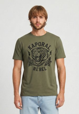 Młody mężczyzna z jasnobrązowymi włosami, ubrany w oliwkowo-zielony t-shirt z ryczącym tygrysem i napisem „Kaporal Rebel”, stoi na jednolitym tle.