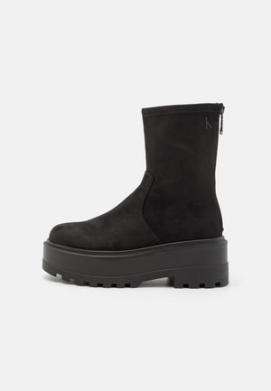 Musta nilkkuri, jossa on paksu urheilupohja, sivuvetoketju ja hienovarainen Calvin Kleinin logo painettuna ylös ja pohjaan.