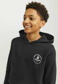 Črna kapuca z sprednjim kenguru žepom in okroglo belo logo, ki vsebuje besedilo in akcent. Tkanina se zdi mehka in strukturirana.