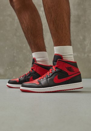 Nogi w czarno-czerwonych butach Air Jordan 1 z białymi skarpetkami, stojące na szarej podłodze przy szarej teksturowanej ścianie.