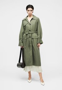 Μακρύ χακί μουσταρδί παλτό τύπου τραντσκότ με ζώνη στη μέση, μεγάλο γιακά και κούμπωμα με κουμπιά. Διαθέτει δύο τσέπες μπροστά και μακριά μανίκια.