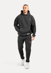 Set di felpa con cappuccio e pantaloni tuta neri realizzato in tessuto morbido. Presenta una tasca a marsupio, etichette con numeri e sneaker bianche per un look informale.