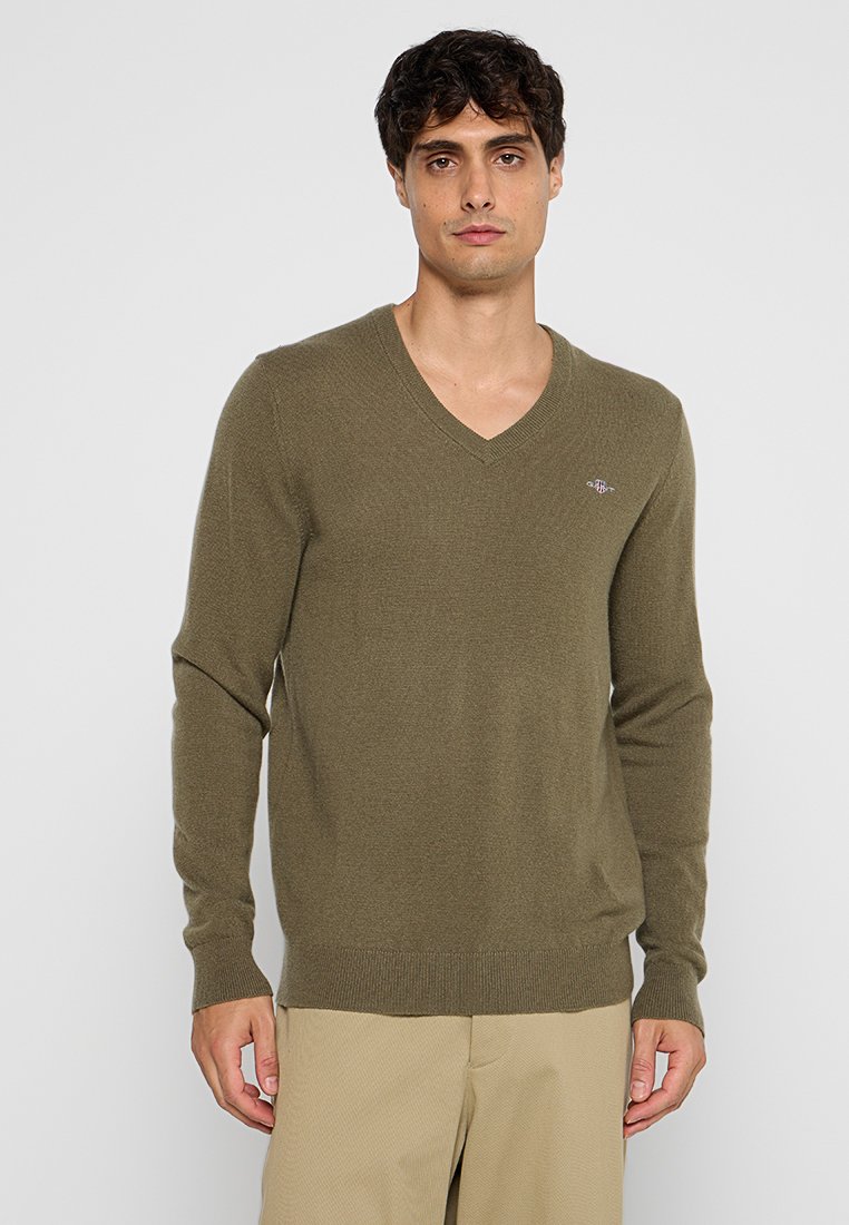 Pull en V couleur olive. Texture tricotée, manches longues, coupe ajustée. Petit logo sur la poitrine, poignets et ourlet contrastants.