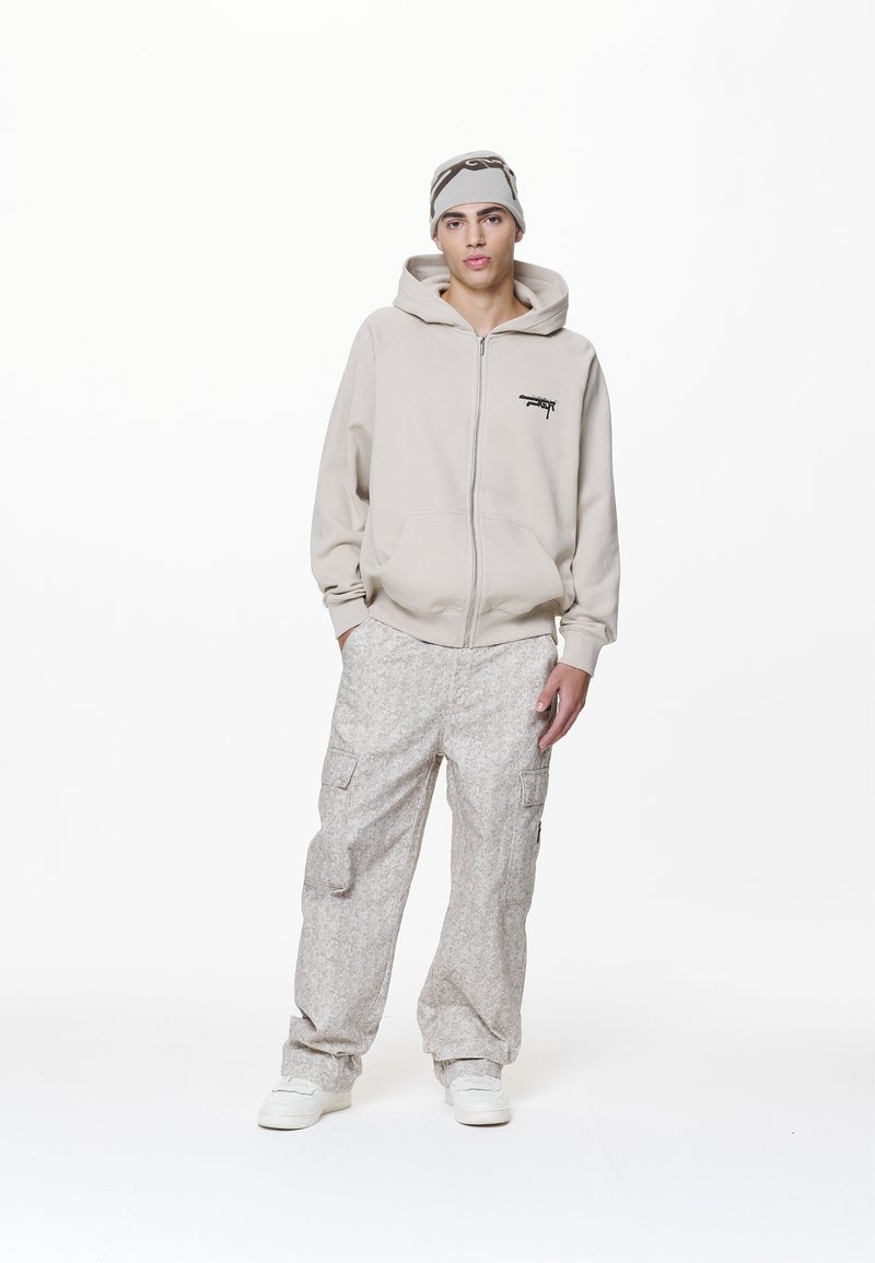 Beige Zip-Hoodie mit Fronttaschen, kombiniert mit hellgrauen gemusterten Cargo-Hosen. Das Model trägt eine passende Mütze und weiße Sneakers.