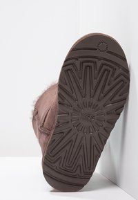 Brązowy zamszowy but z teksturowaną gumową podeszwą, zdobiony wzorem promieni słonecznych oraz wytłoczonym logo. Zawiera miękkie futro na górze.