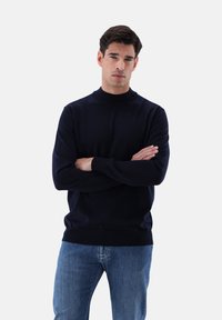 Marineblauer Strickpullover mit hohem Kragen und langen Ärmeln, der über eine glatte Textur sowie gerippte Bündchen und Saum verfügt. Getragen mit blauen Jeans.