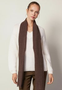 Foulard marron en tissu doux et texturé, drapé légèrement autour du cou, associé à un pull en tricot blanc et à un pantalon olive.