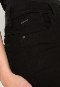 Acercamiento del bolsillo de unos pantalones vaqueros G-Star negros con la etiqueta de marca visible y una mano parcialmente visible descansando cerca de la cintura.