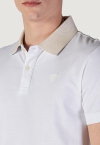 Camisa polo blanca con un cuello de punto beige, que cuenta con una tapeta de botones y un sutil logo triangular en el lado izquierdo del pecho. Textura de tela suave.