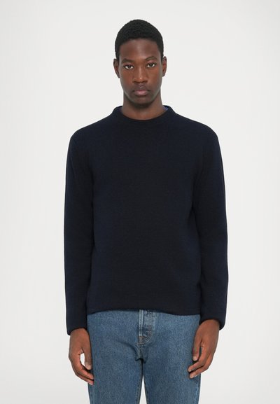 Donker marineblauw crew neck sweater van zacht breimateriaal met lange mouwen, gedragen met lichtblauwe denim jeans. Eenvoudige, klassieke pasvorm.