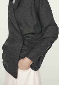 Ciemnoszara, teksturowana wełniana kurtka o luźnym kroju, z pojedynczym czarnym guzikiem i boczną kieszenią, noszona na jasnym ubraniu.