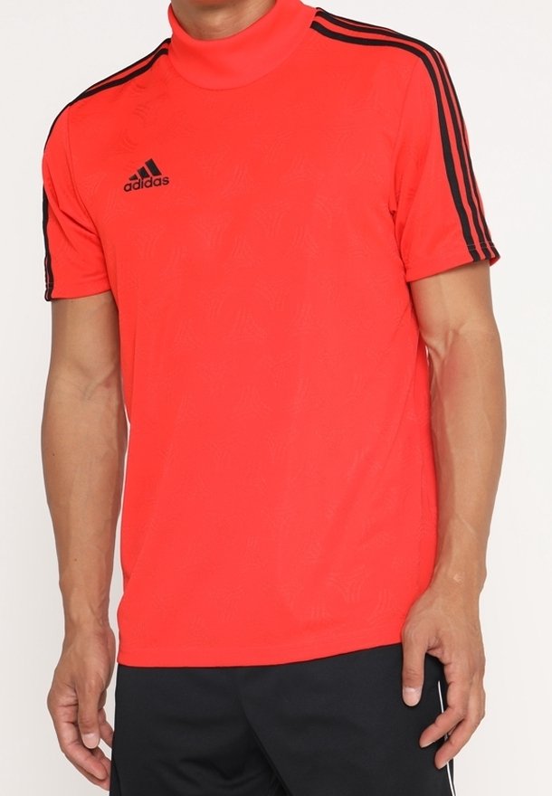 Mies, joka pukeutuu kirkkaan punaiseen, mustilla olkapääraidoilla ja mustalla Adidas-logolla rinnassa olevaan lyhythihaiseen Adidas-urheilupaitaan.