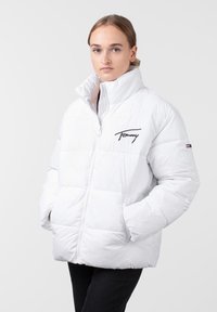 Bijela puffer jakna s visokim ovratnikom, zatvaranjem na zip i izvezеним "Tommy" logom na prsima. Ima kvadratni dizajn i bočne džepove.