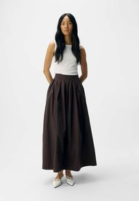 Gonna maxi a pieghe marrone con vita alta, abbinata a un top a canotta bianco aderente e a delle ballerine bianche a punta. Tessuto liscio, silhouette lunga.